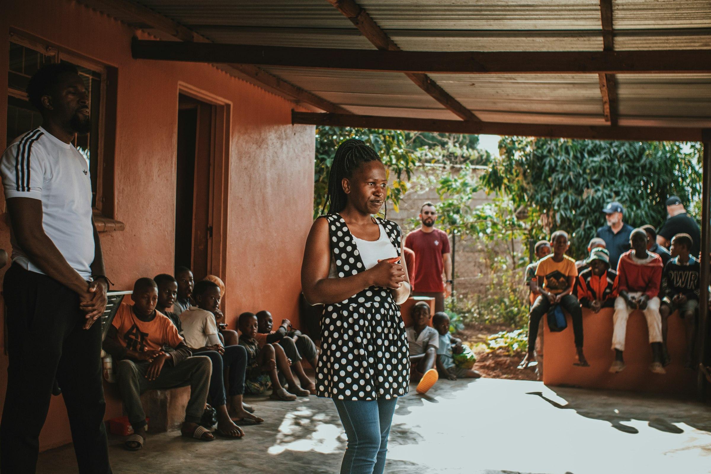 A woman speaking to a seated gathering under a corrugated roof.