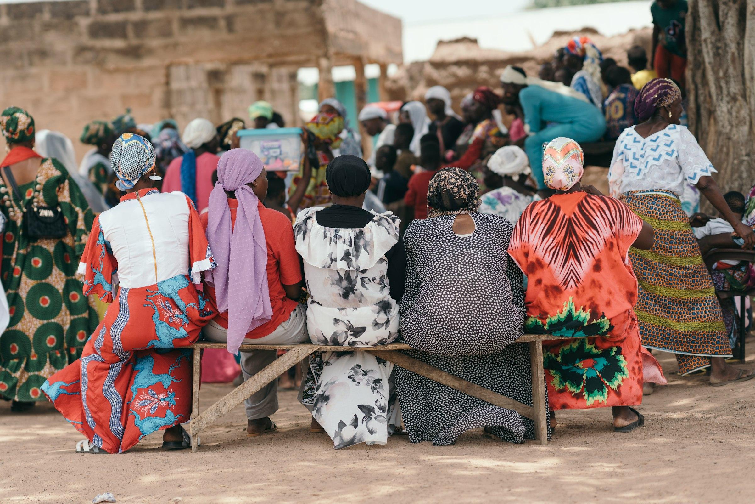 Women in patterned dress seated on a bench with their backs to the camera, facing a village gathering.