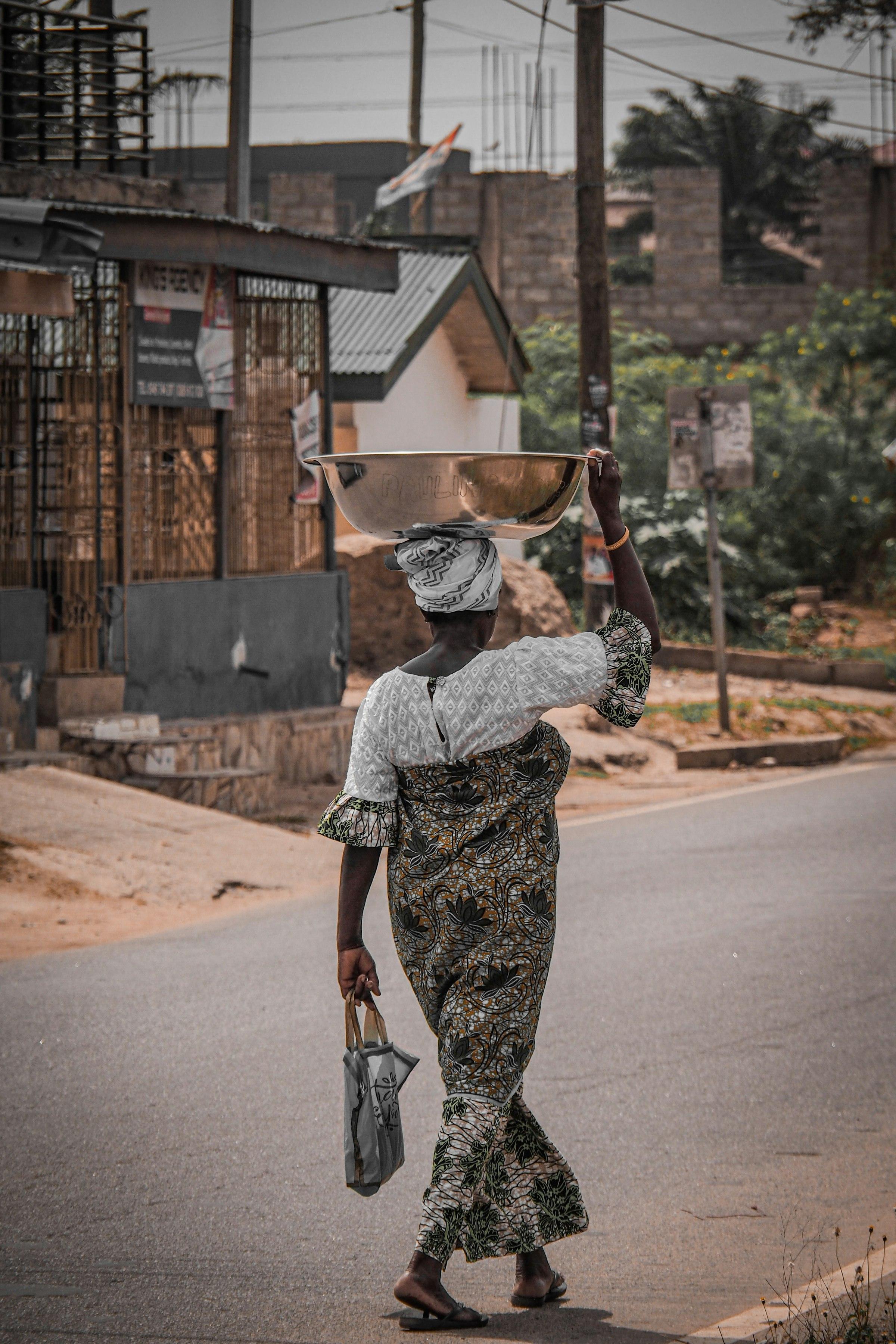 A woman crossing a street with a metal basin balanced on her head, a roadside shopfront behind her.