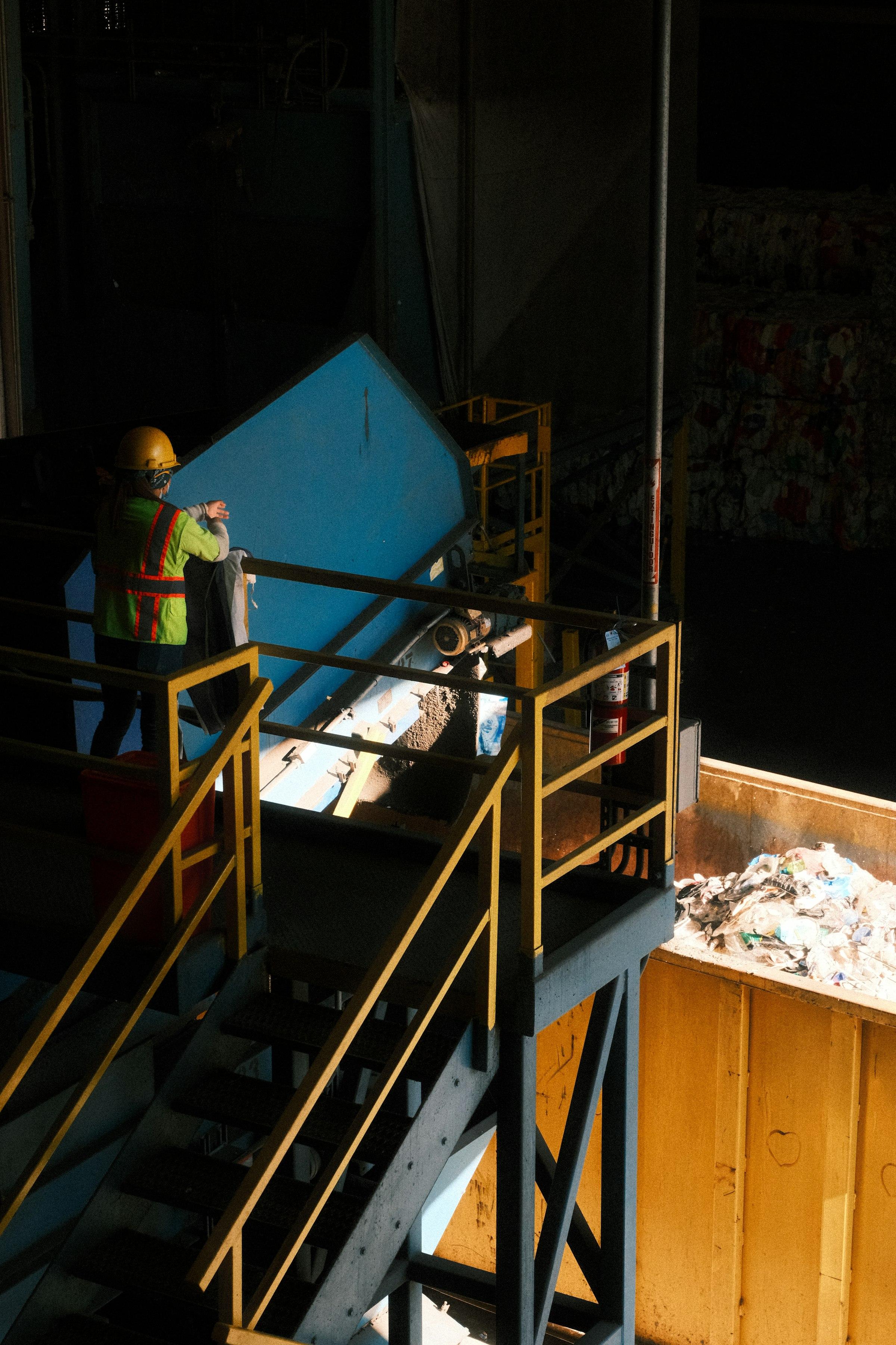 A worker in a hard hat and high-visibility vest on a gantry above a waste sorting line, lit by a single shaft of daylight.
